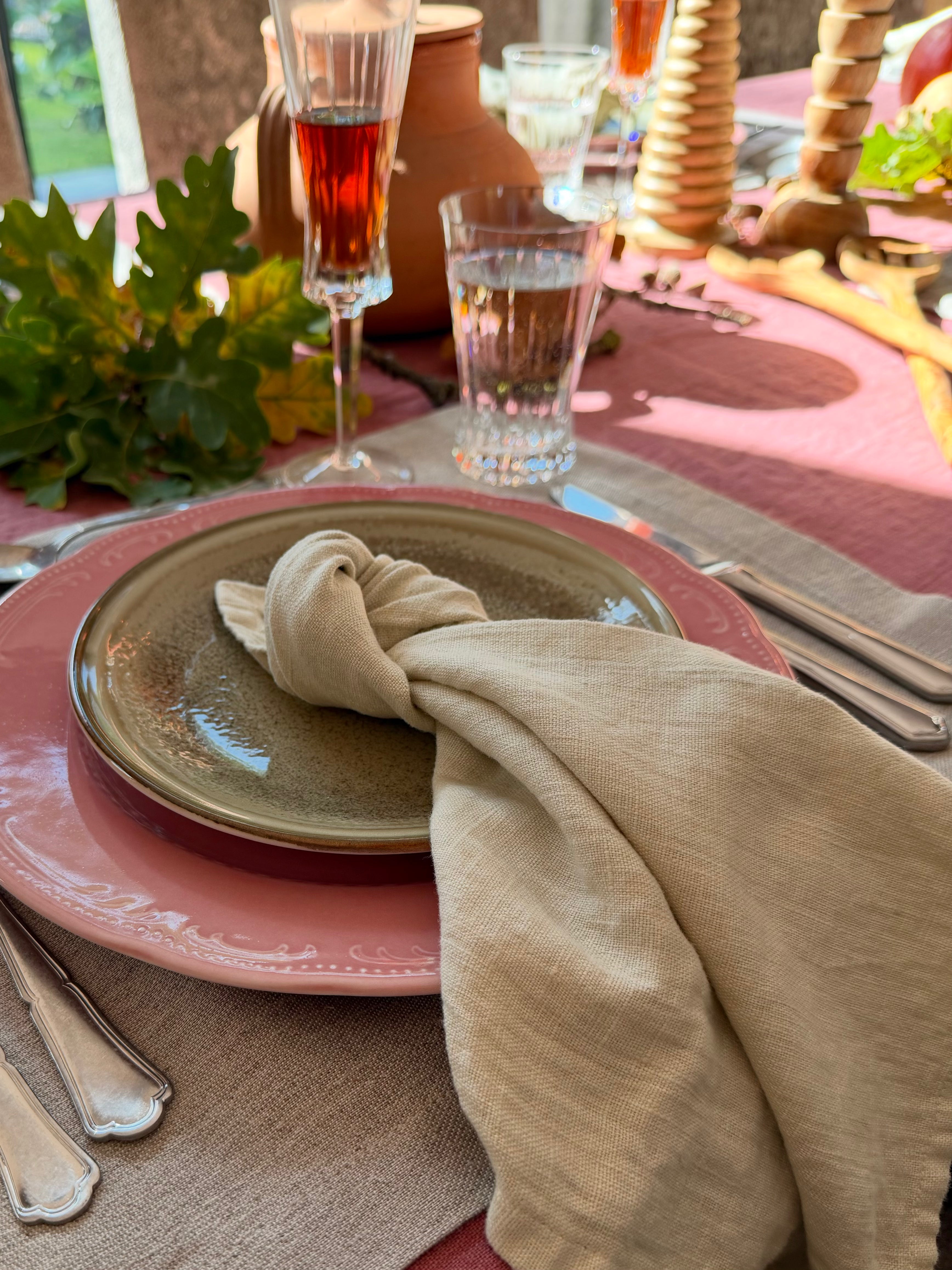 Sfeervol gedekte herfsttafel met oudroze tafellinnen, naturel linnen placemat en servet, gecombineerd met roze en aardewerken servies. Glazen, zilverkleurig bestek, herfstbladeren en houten kandelaars zorgen voor een warme, Toscaanse sfeer.