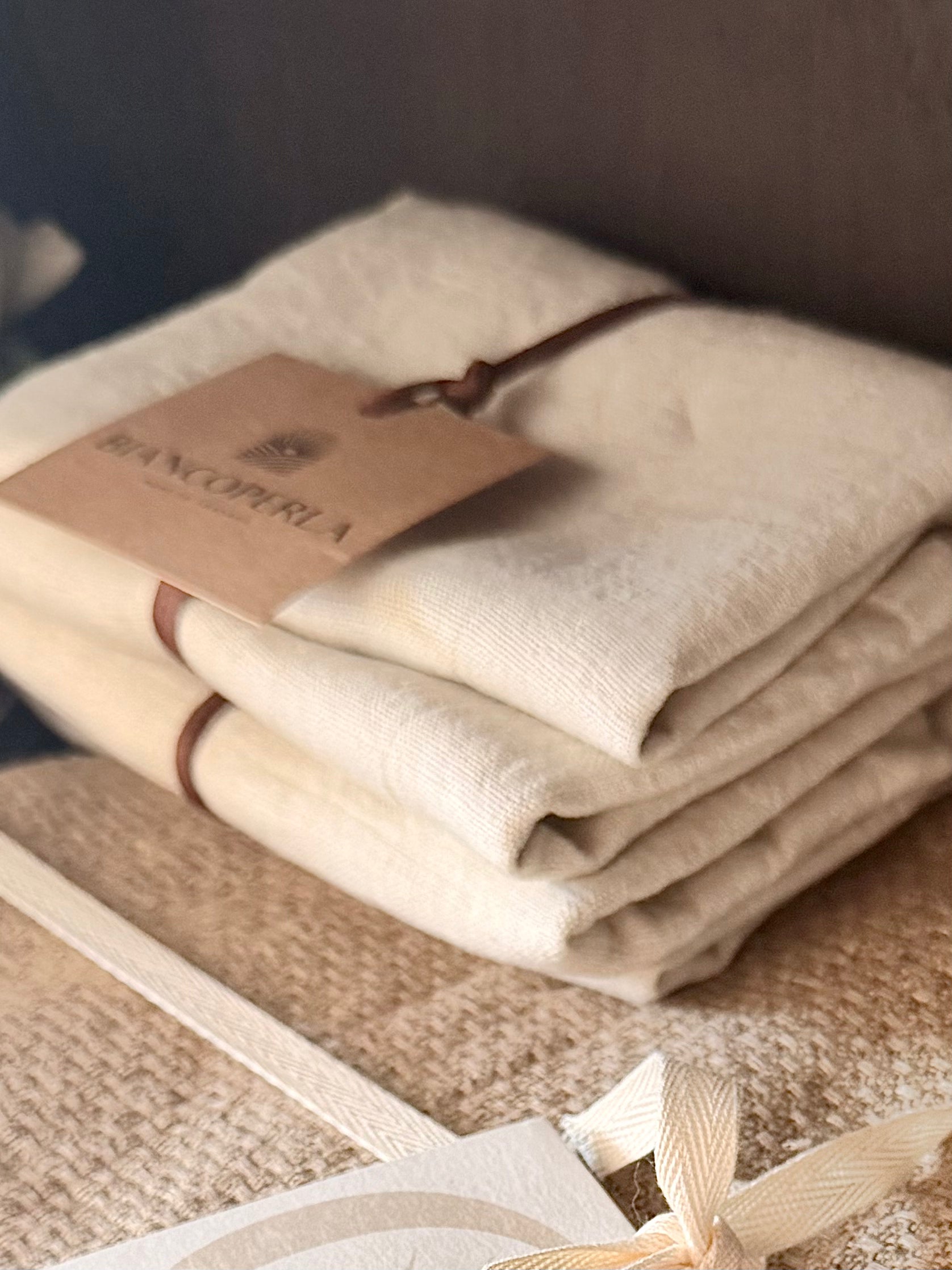 Detailfoto van een stapel naturel linnen servetten van Biancoperla, afgewerkt met een leren koord en merklabel, gecombineerd met grof geweven tafellinnen in een warme natureltint.